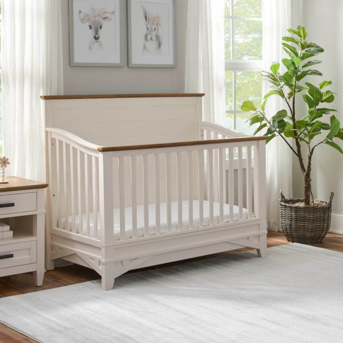 A white Heritage House Artisan 4-in-1 convertible crib with a wooden frame next to a matching wooden dresser.